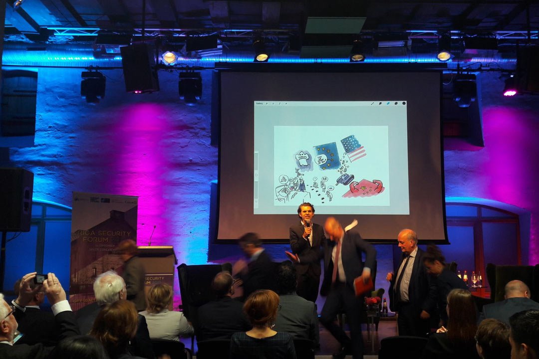 Fonā uz liela ekrāna redzama karikatūra, kurā fiksēts iepriekšējā konferences diskusijā runātais. Viens no klātesošajiem fotografē karikatūru ar savu viedtālruni.