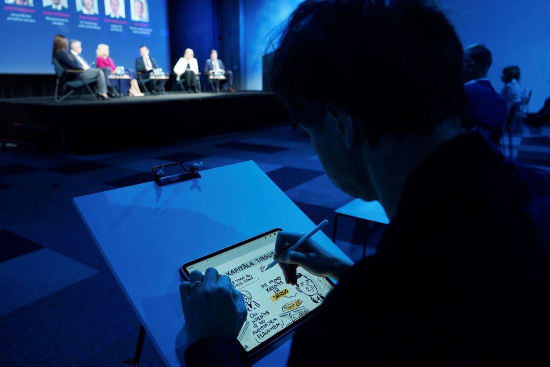 Uz skatuves konferencē notiek diskusija un karikatūrists tajā pārrunāto vizualizē zīmētā veidā uz digitālās planšetes